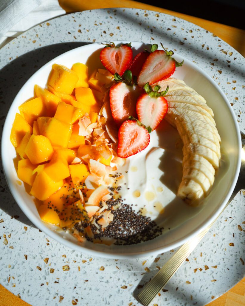Zoomed-out shot of a white bistro bowl filled with a tropical mango strawberry Greek yogurt bowl, set on a circular white terrazzo riser on a sunlit table.