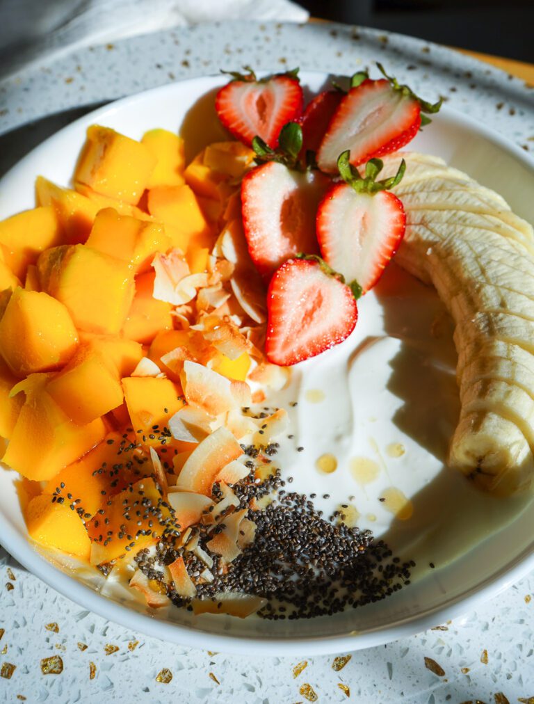 A white bistro bowl filled with creamy Greek yogurt, diced fresh mango, halved strawberries, banana slices, toasted coconut flakes, and a honey drizzle.