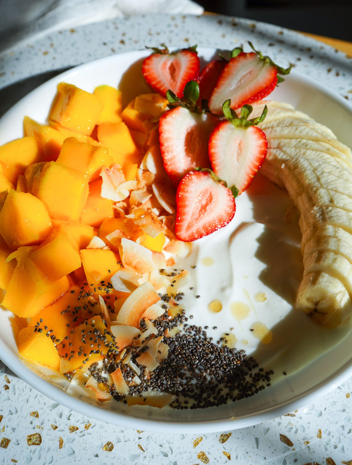 A white bistro bowl filled with creamy Greek yogurt, diced fresh mango, halved strawberries, banana slices, toasted coconut flakes, and a honey drizzle.