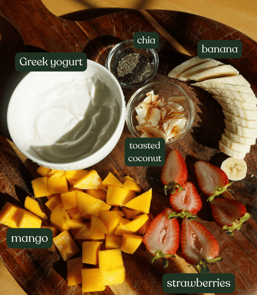 Flat-lay image of yogurt bowl ingredients on a wooden board labeled with dark green badges for Greek yogurt, mango, strawberries, banana, chia, and toasted coconut.