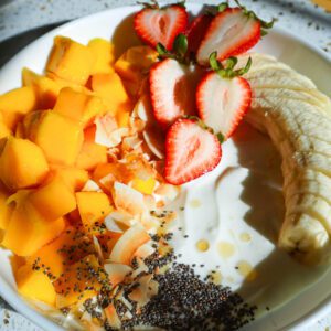 Square overhead crop of a full white bistro bowl filled with Greek yogurt, diced mango, fresh strawberries, banana slices, chia seeds, and toasted coconut.